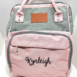 Pu&ograve; includere: Una borsa per pannolini rosa e grigia con una toppa marrone che presenta un uccello dei cartoni animati e il testo "KeaBabies". La borsa ha una tasca con cerniera sul davanti con il nome "Kynleigh" ricamato in nero.