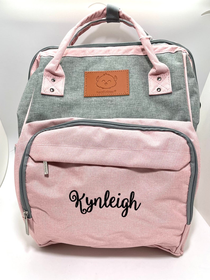 Pu&ograve; includere: Una borsa per pannolini rosa e grigia con una toppa marrone che presenta un uccello dei cartoni animati e il testo "KeaBabies". La borsa ha una tasca con cerniera sul davanti con il nome "Kynleigh" ricamato in nero.