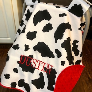 May include: A white and black cow print car seat cover with a red trim and a red plush backing. The cover has the name "DUSTIN" embroidered in red on the front.