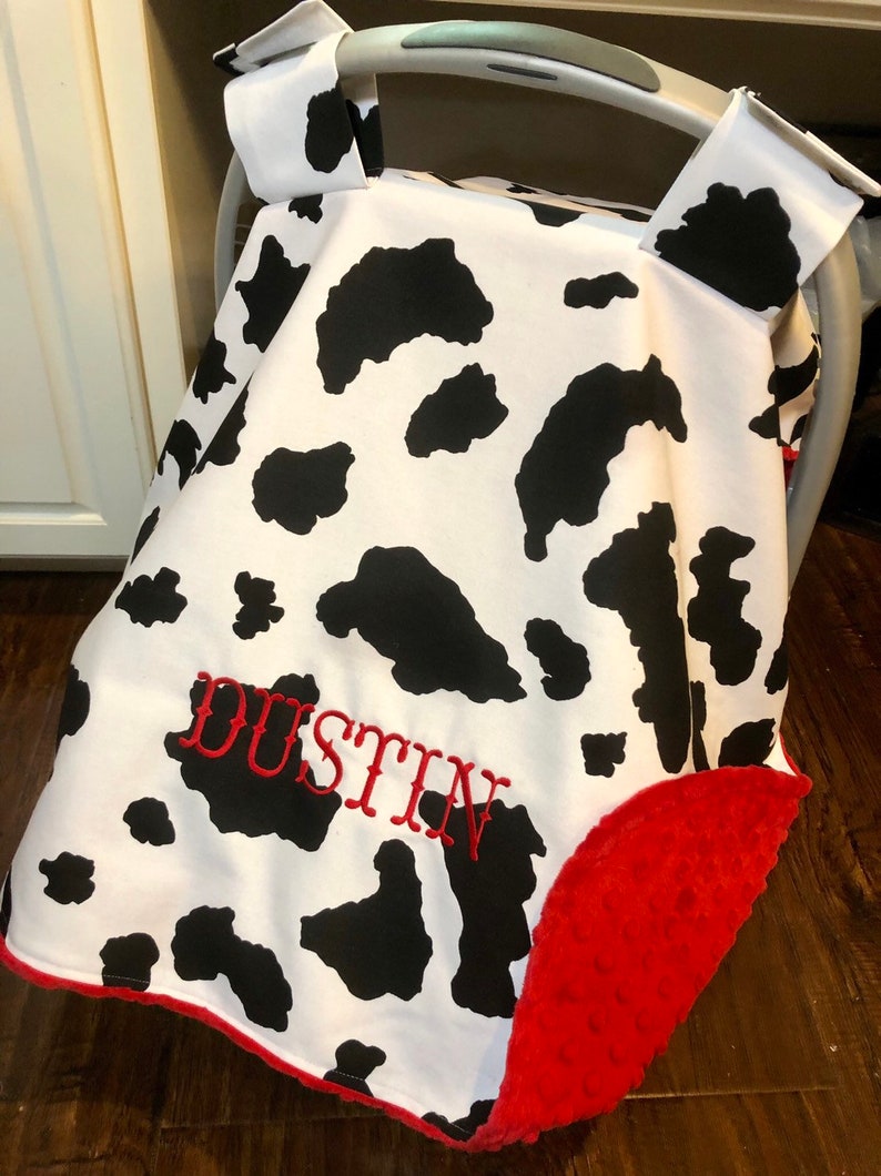 May include: A white and black cow print car seat cover with a red trim and a red plush backing. The cover has the name "DUSTIN" embroidered in red on the front.