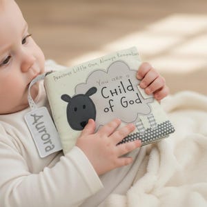May include: A soft baby book with a sheep design and the text "You are a Child of God." The book has a tag that says "Aurora." The book is cream-colored with gray accents and is being held by a baby.