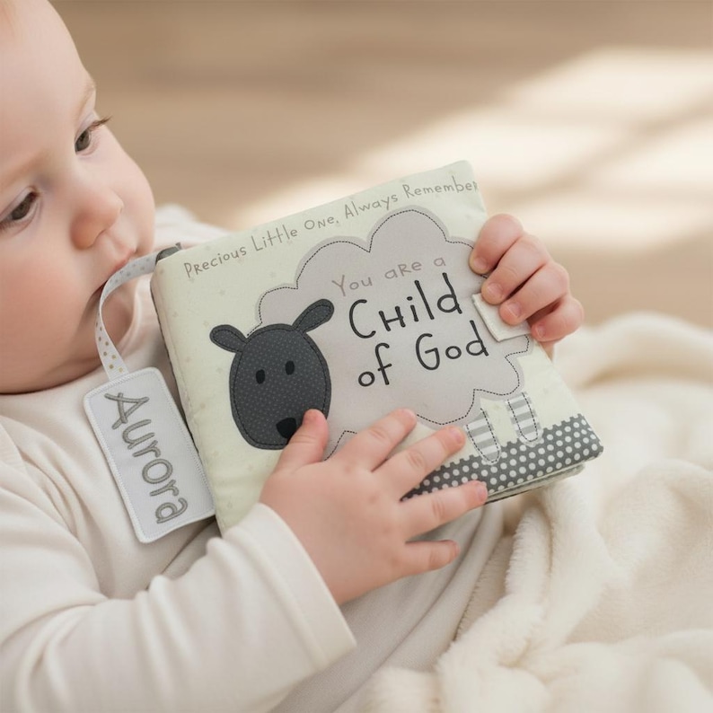 May include: A soft baby book with a sheep design and the text "You are a Child of God." The book has a tag that says "Aurora." The book is cream-colored with gray accents and is being held by a baby.
