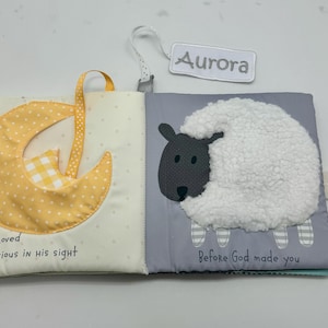 May include: Open baby book with a yellow dotted moon and a white sheep with a gray face. The text "Dearly Loved and precious in his sight" and "Before God made you" are printed on the pages. A tag that says "Aurora" is attached.