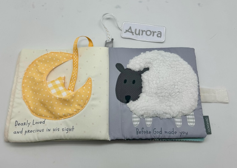 May include: Open baby book with a yellow dotted moon and a white sheep with a gray face. The text "Dearly Loved and precious in his sight" and "Before God made you" are printed on the pages. A tag that says "Aurora" is attached.