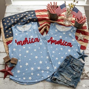 May include: Two light blue sleeveless shirts featuring a white star pattern. One shirt has the red text 'merica and the other has the red text America. The shirts are displayed with denim shorts, small American flags, and wooden blocks marked 1776.