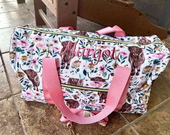 May include: A white duffel bag featuring a repeating pattern of brown Highland cattle and pink and brown flowers. The name Margot is embroidered in pink script on the front. The bag includes pink nylon handles and a gold-toned zipper closure.