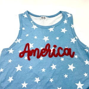 May include: A light blue sleeveless tank top featuring a white star pattern throughout the fabric. The word America is written across the chest in a red, textured, cursive-style applique. A white label with black text is visible at the neckline.