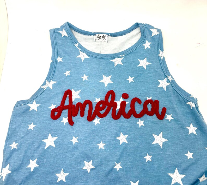 May include: A light blue sleeveless tank top featuring a white star pattern throughout the fabric. The word America is written across the chest in a red, textured, cursive-style applique. A white label with black text is visible at the neckline.