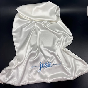 May include: A white satin bonnet featuring a smooth, reflective surface and a drawstring closure. The name "josie" is embroidered in blue cursive script across the lower center of the fabric, with a small blue heart above the letter i.