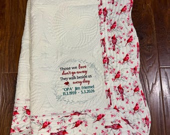 May include: A cream-colored quilted throw blanket featuring a central panel with embroidered text that reads: Those we love don't go away They walk beside us every day 'OPA' Jim Friemel 11.3.1959 - 5.3.2026, bordered by a red cardinal bird pattern.