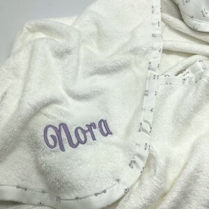 May include: A white hooded baby towel with the name "Nora" embroidered in lavender thread. The towel has a patterned border with small paw prints and bones. A matching bib is visible in the background.