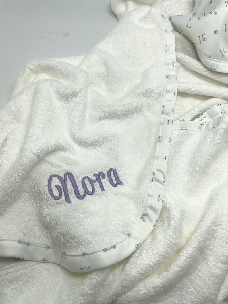 May include: A white hooded baby towel with the name "Nora" embroidered in lavender thread. The towel has a patterned border with small paw prints and bones. A matching bib is visible in the background.