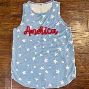 May include: A light blue sleeveless tank top featuring a repeating pattern of white stars across the fabric. The word America is embroidered in red cursive script across the center of the chest. The garment has a round neckline and a curved hem.