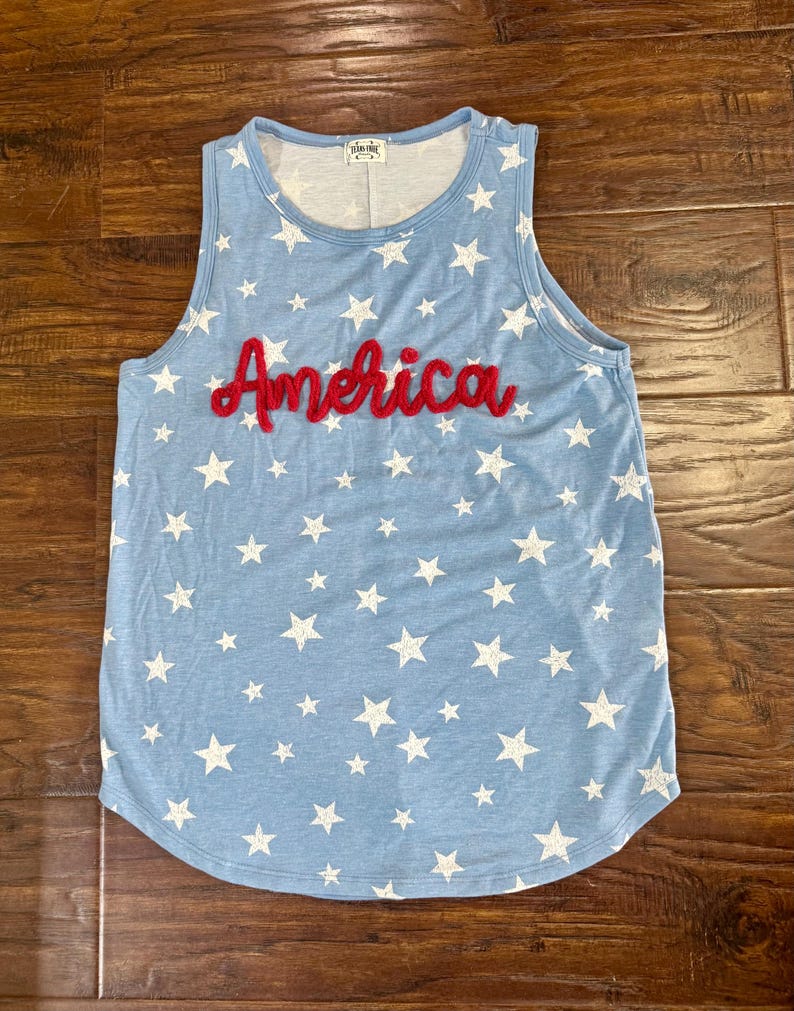 May include: A light blue sleeveless tank top featuring a repeating pattern of white stars across the fabric. The word America is embroidered in red cursive script across the center of the chest. The garment has a round neckline and a curved hem.