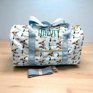 May include: A light blue duffel bag featuring a duck pattern and the name "HARVEY" embroidered in green. The bag has light blue handles, a detachable shoulder strap, and gold-coloured zips. The duck pattern shows ducks in flight and swimming.