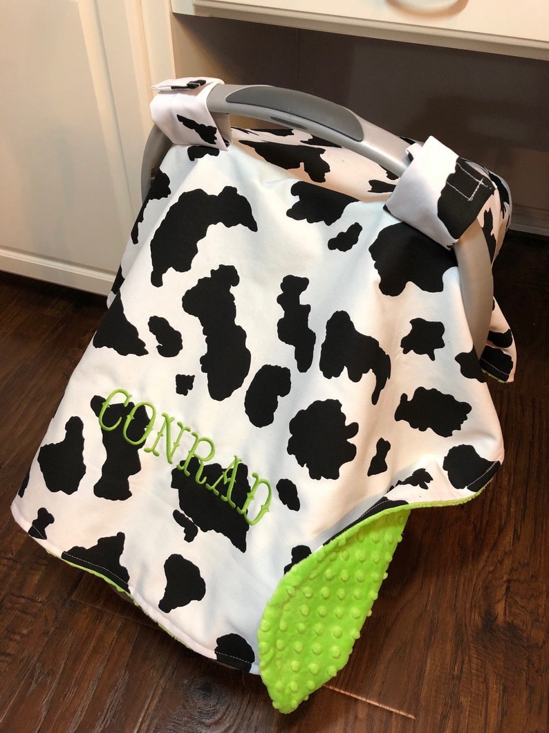 May include: A black and white cow print car seat cover with a green and white plush trim. The cover has the name "CONRAD" embroidered on it.