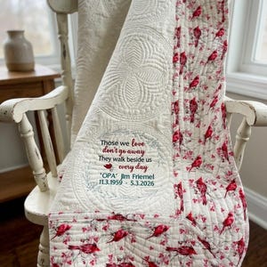 May include: A cream-colored quilted throw blanket featuring a floral and cardinal bird pattern border. The center panel displays embroidered text: Those we love don't go away They walk beside us every day 'OPA' Jim Friemel 11.3.1959 - 5.3.2026.