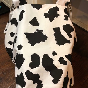May include: A white and black cow print car seat cover with green trim. The cover is made of soft fabric and is designed to protect your car seat from spills and stains.