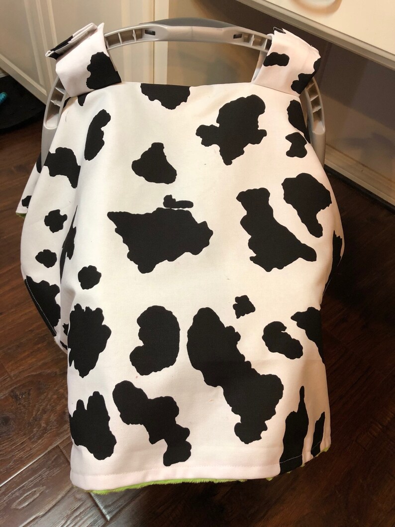 May include: A white and black cow print car seat cover with green trim. The cover is made of soft fabric and is designed to protect your car seat from spills and stains.