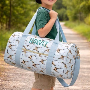 May include: A light blue duffel bag with a duck pattern and the name "HARVEY" in green. The bag has light blue straps and a gold zip. The bag is being carried by a person wearing a green top, khaki shorts, and a navy blue baseball cap.
