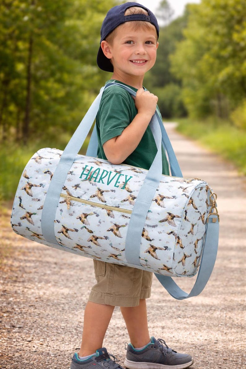 May include: A light blue duffel bag with a duck pattern and the name "HARVEY" in green. The bag has light blue straps and a gold zip. The bag is being carried by a person wearing a green top, khaki shorts, and a navy blue baseball cap.