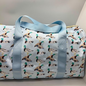 May include: A light blue duffel bag featuring a pattern of mallard ducks. The bag has light blue handles and a zip closure. The bag is approximately 30 cm high and 61 cm long.