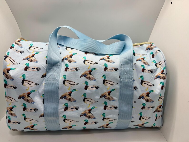 May include: A light blue duffel bag featuring a pattern of mallard ducks. The bag has light blue handles and a zip closure. The bag is approximately 30 cm high and 61 cm long.
