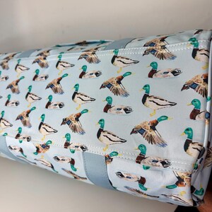 May include: A rectangular tote bag featuring a light blue background adorned with a repeating pattern of mallard ducks. The ducks are shown in various poses, with green heads, brown wings, and white and black bodies. The bag has light blue handles.