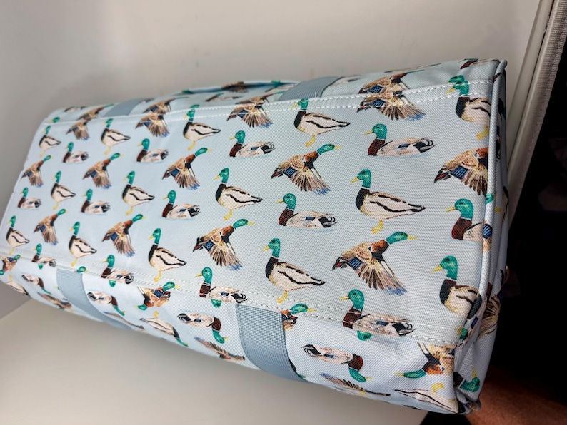 May include: A rectangular tote bag featuring a light blue background adorned with a repeating pattern of mallard ducks. The ducks are shown in various poses, with green heads, brown wings, and white and black bodies. The bag has light blue handles.