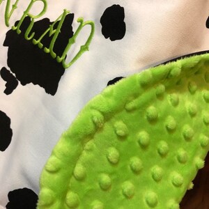 May include: A green and white baby blanket with a cow print pattern and the word "WRAN" embroidered on it. The blanket has a soft, textured side with small raised dots.