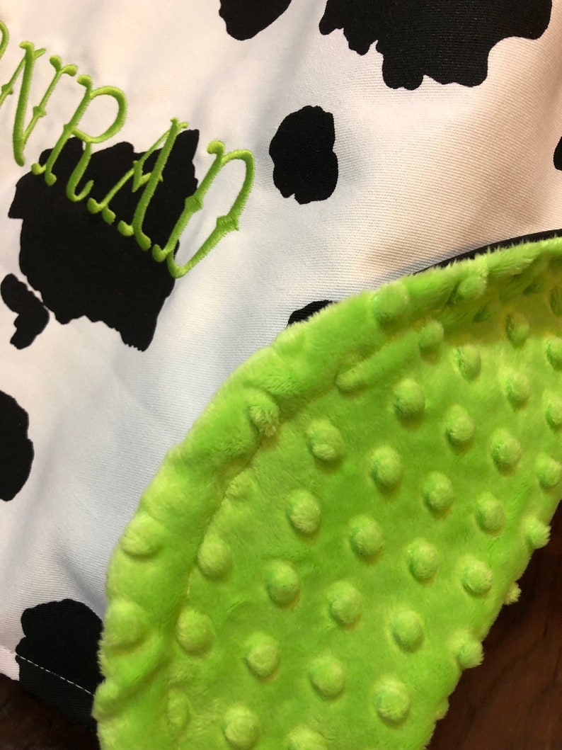 May include: A green and white baby blanket with a cow print pattern and the word "WRAN" embroidered on it. The blanket has a soft, textured side with small raised dots.