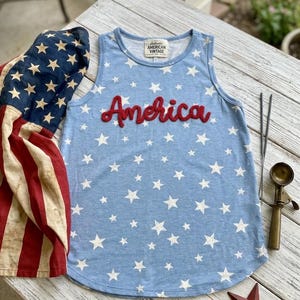 May include: A light blue sleeveless tank top featuring a white star pattern and the word America in red script lettering across the chest. The garment has a label reading Authentic American Vintage at the inner neckline and a rounded hemline.
