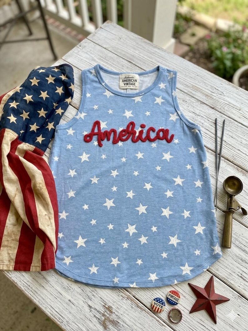 May include: A light blue sleeveless tank top featuring a white star pattern and the word America in red script lettering across the chest. The garment has a label reading Authentic American Vintage at the inner neckline and a rounded hemline.
