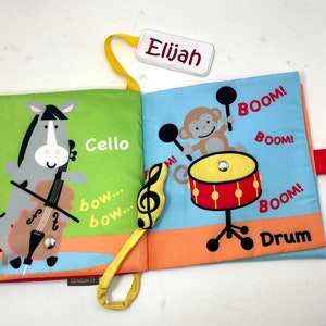 May include: A colorful cloth book for babies and toddlers with a grey horse playing a cello and a monkey playing a drum. The book has the words "Cello", "bow... bow...", "BOOM!" and "Drum" printed on it. The book is personalized with the name "Elijah".