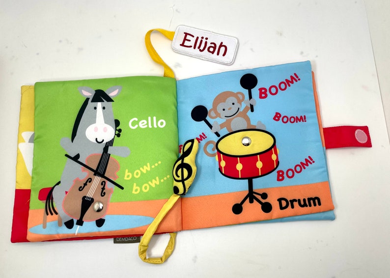 May include: A colorful cloth book for babies and toddlers with a grey horse playing a cello and a monkey playing a drum. The book has the words "Cello", "bow... bow...", "BOOM!" and "Drum" printed on it. The book is personalized with the name "Elijah".