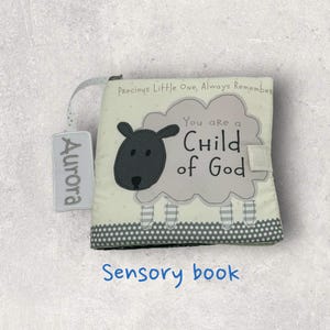 May include: A soft sensory book with a sheep design. The book features the text "You are a Child of God" and "Precious Little One, Always Remember". A tag with the name "Aurora" is attached. The bottom of the book has a gray polka dot pattern.