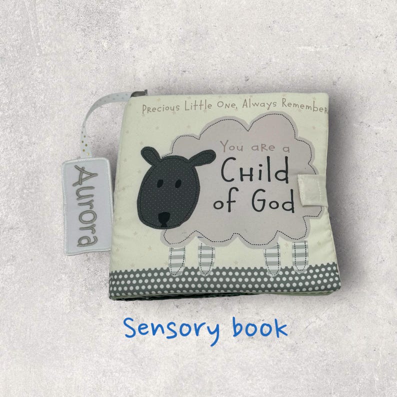 May include: A soft sensory book with a sheep design. The book features the text "You are a Child of God" and "Precious Little One, Always Remember". A tag with the name "Aurora" is attached. The bottom of the book has a gray polka dot pattern.