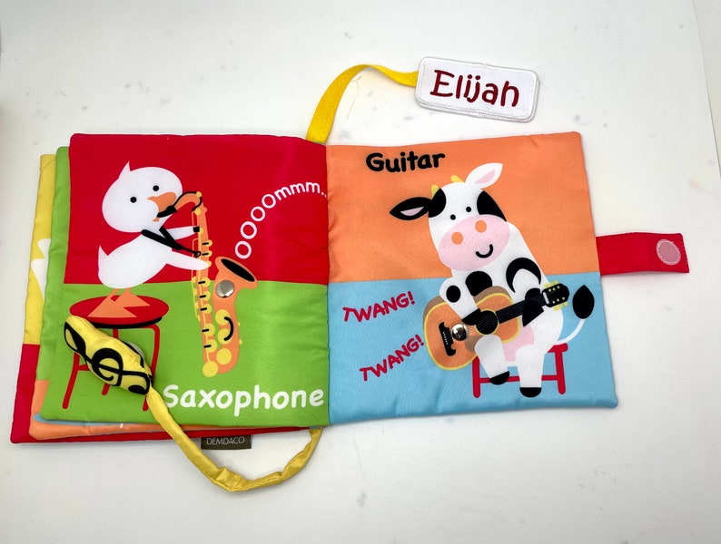 May include: A colorful cloth book for babies and toddlers with a duck playing a saxophone and a cow playing a guitar. The book has the words "Saxophone", "Oommm", "Guitar", "Twang!" and "Elijah" on it.