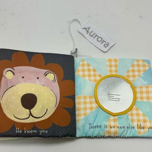 May include: Open soft baby book with a lion illustration on one page and a mirror on the other. The lion is brown, pink, and yellow. The mirror is surrounded by a yellow sunburst design. Text reads "He knew you" and "There is no one else like you."