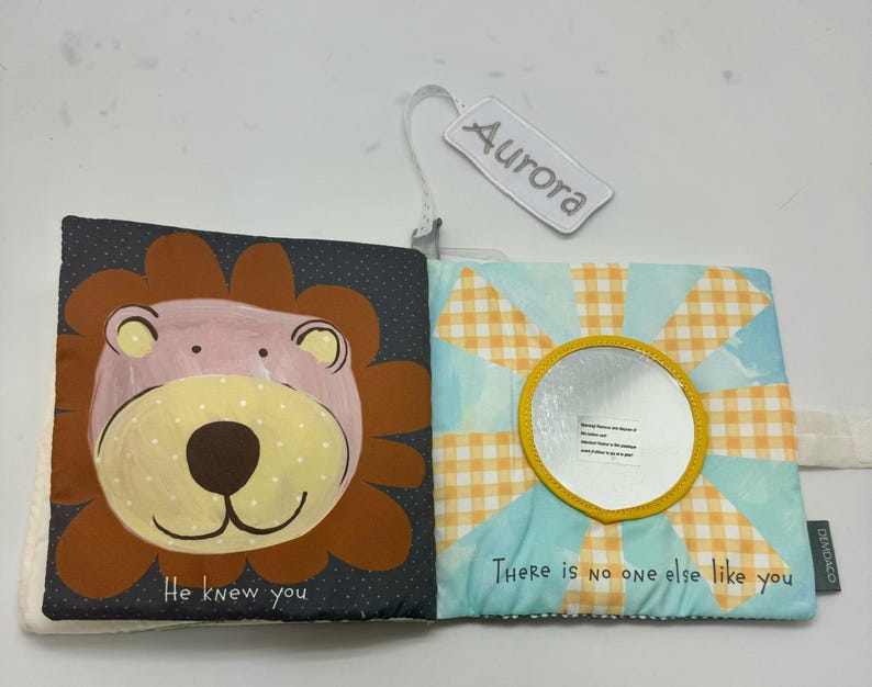 May include: Open soft baby book with a lion illustration on one page and a mirror on the other. The lion is brown, pink, and yellow. The mirror is surrounded by a yellow sunburst design. Text reads "He knew you" and "There is no one else like you."