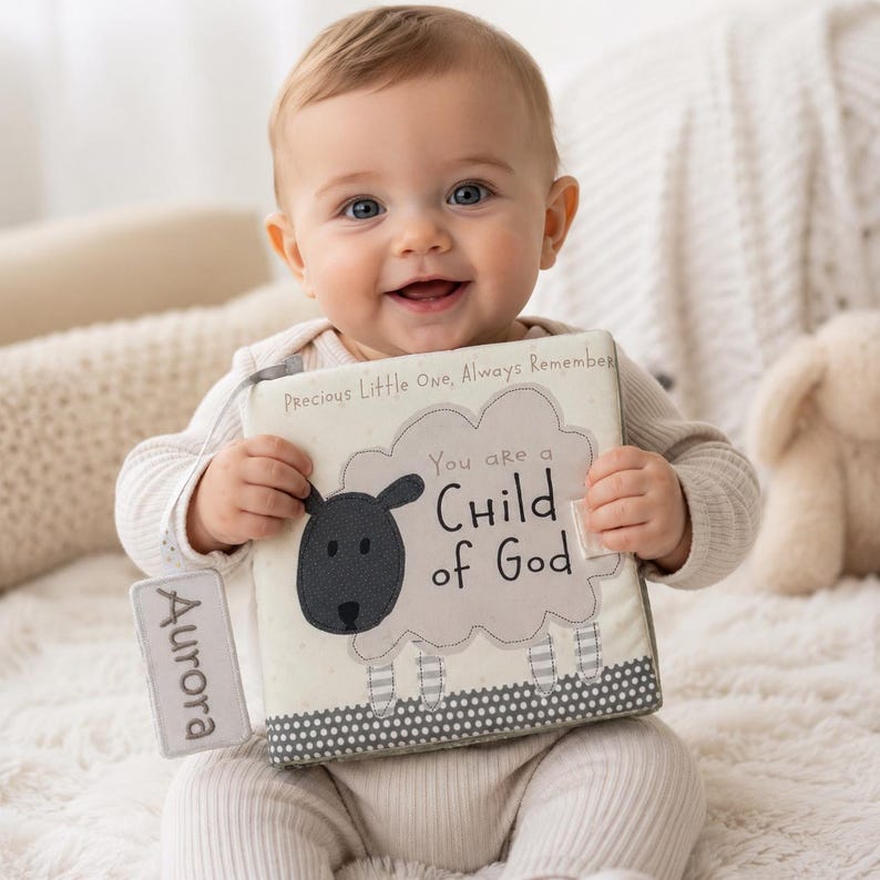 May include: A baby holding a soft, cloth book with the text "You are a Child of God." The book features a sheep design and the name "Aurora" on a tag. The book has a gray and white border.