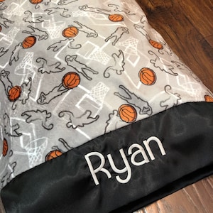 May include: A gray and white fleece pillow with a basketball theme. The pillow has a black satin border with the name "Ryan" embroidered in white.