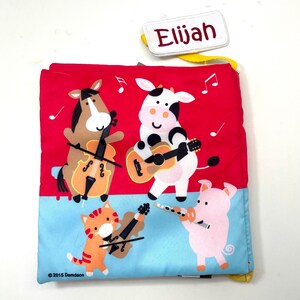 May include: A red and blue fabric book with a personalized name tag that says "Elijah". The book features a cow playing a guitar, a horse playing a violin, a cat playing a violin, and a pig playing a flute. The book is titled "2015 Demdaco".