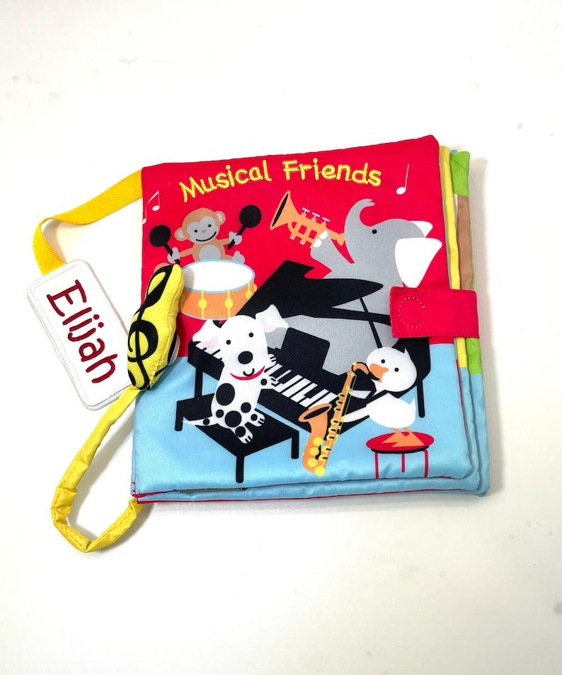 May include: A red, yellow, blue, and green fabric book with the title "Musical Friends" and a name tag that says "Elijah". The book features a monkey playing drums, a dog playing piano, an elephant playing trumpet, and a duck playing saxophone.