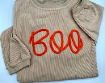 May include: A beige long-sleeve shirt made of ribbed fabric featuring the word BOO written in bright orange fuzzy textured lettering across the chest. The garment has a round neckline and ribbed cuffs on the sleeves, providing a consistent texture.
