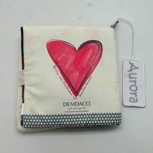 May include: A soft, cloth baby book with a large red heart illustration on the cover. The book is titled "Aurora" and includes the text "Written and Illustrated by Jennifer Heynen" and "DEMDACO. lift the spirit."