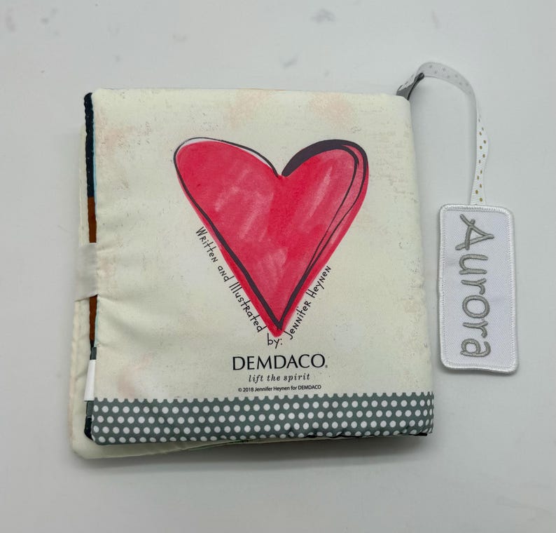 May include: A soft, cloth baby book with a large red heart illustration on the cover. The book is titled "Aurora" and includes the text "Written and Illustrated by Jennifer Heynen" and "DEMDACO. lift the spirit."