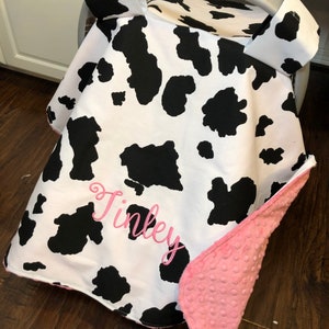 May include: A white and black cow print car seat cover with a pink fleece lining. The cover has a pink embroidered name "Finley" on the front.