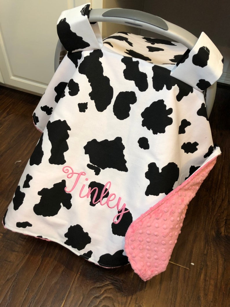 May include: A white and black cow print car seat cover with a pink fleece lining. The cover has a pink embroidered name "Finley" on the front.