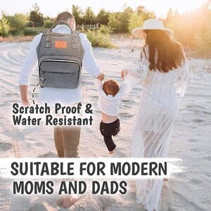 May include: A gray diaper bag backpack with a small bear logo and the text "Scratch Proof & Water Resistant". The bag is being worn by a person walking on a sandy beach with a child and another person.